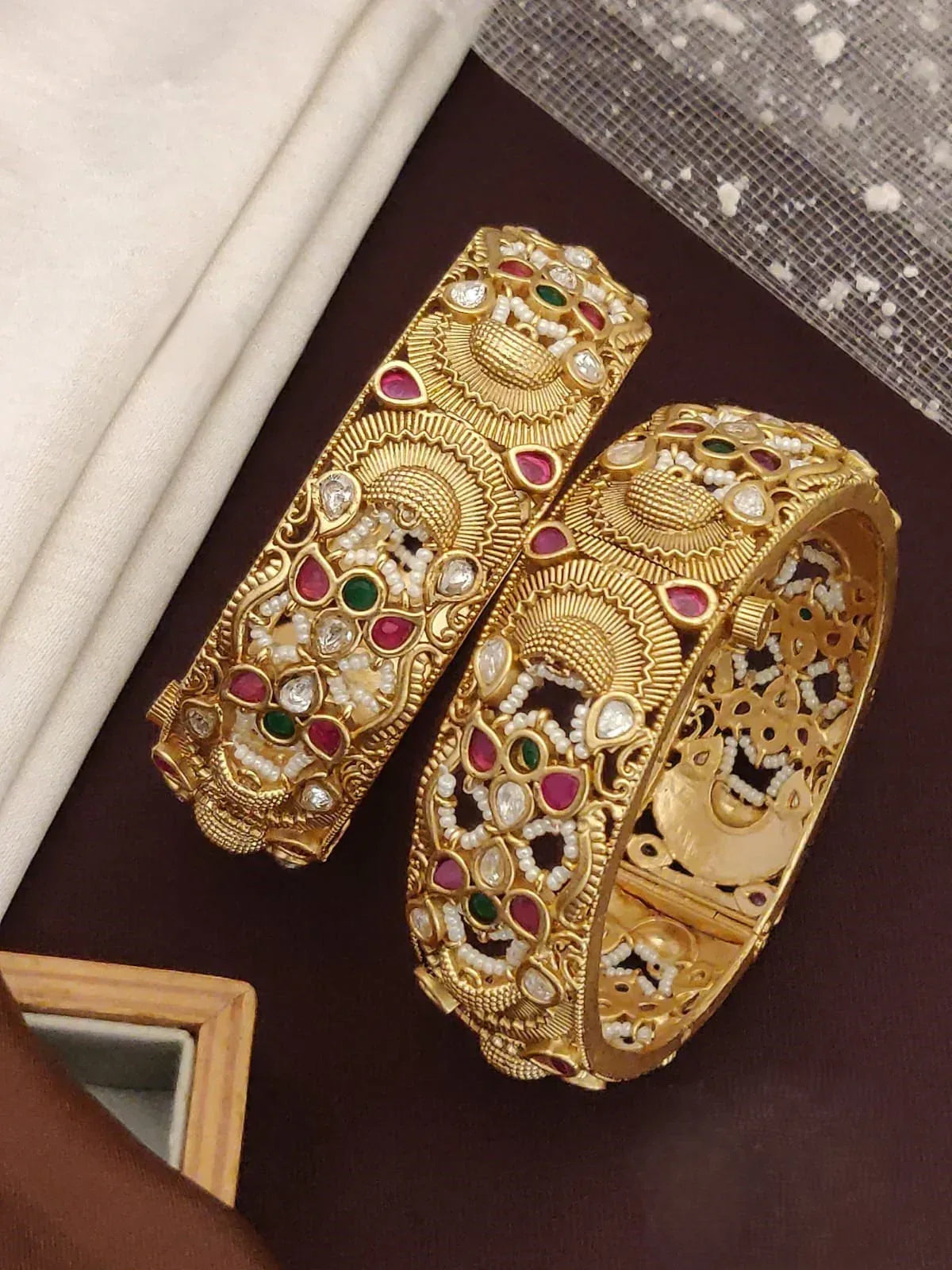 Pair of ornate gold bangles with red, green gemstones and white pearls on dark background