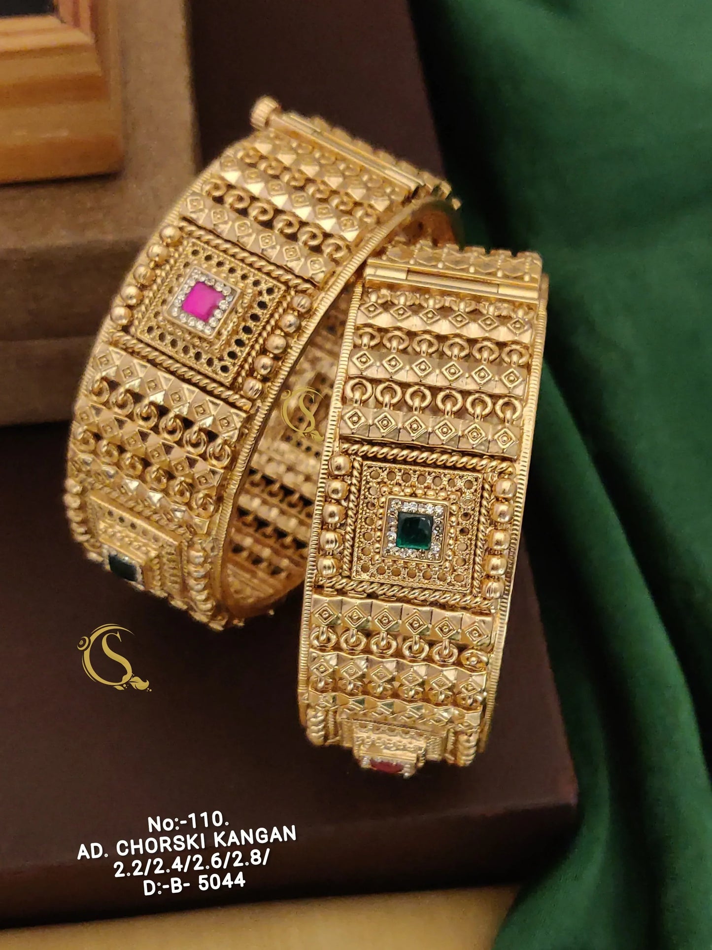Intricately designed gold bangles with detailed patterns and colored square stones, displayed on brown and green fabric