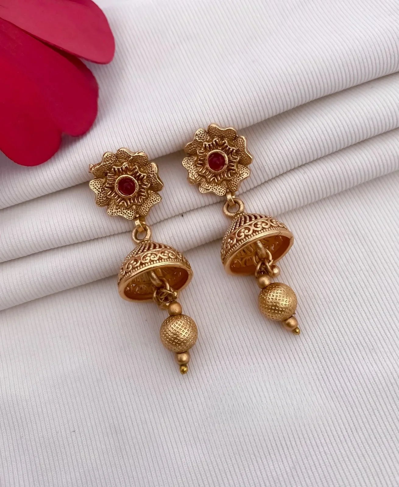Gold-tone floral jhumka earrings with red stone accents on white fabric background