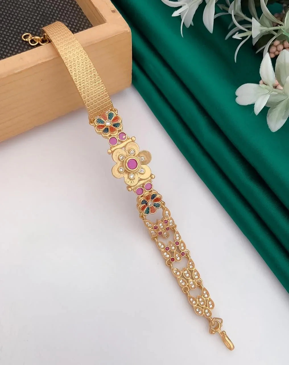 Gold bracelet with pink and multicolor floral designs on white surface with green fabric and white flowers